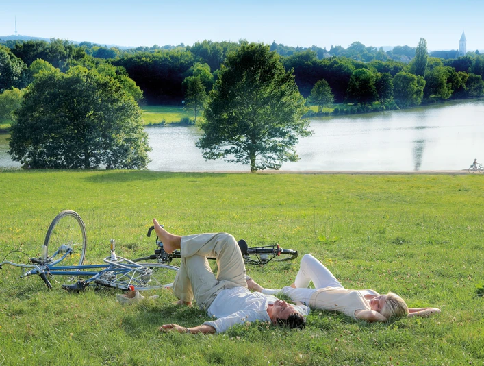 Obersee Bielefeld Zwei Personen liegen entspannt auf einer sonnigen Wiese am Flussufer mit einem Rad im Hintergrund.