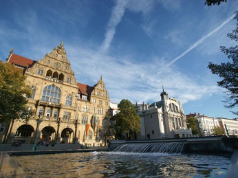Historisches Gebäude und moderner Brunnen im Sonnenschein mit blauem Himmel und Wolkenformationen.