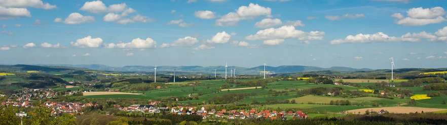 Panoramablick über eine weitläufige grüne Landschaft mit Windrädern und kleinen Siedlungen.