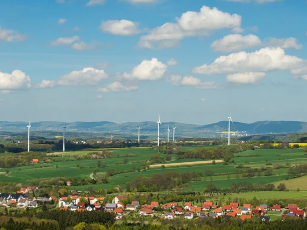 Panoramablick über eine weitläufige grüne Landschaft mit Windrädern und kleinen Siedlungen.