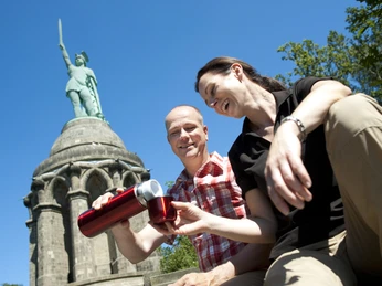 Ein Paar sitzt vor dem Hermannsdenkmal und genießt eine Pause bei strahlend blauem Himmel.