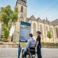 Eine Person im Rollstuhl betrachtet eine Infotafel vor einer Kirche mit gotischem Turm.