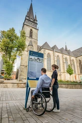 BArrierefreier Stadtrundgang Eine Person im Rollstuhl betrachtet eine Infotafel vor einer Kirche mit gotischem Turm.