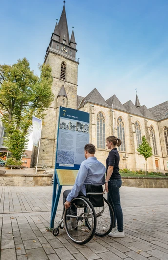 BArrierefreier Stadtrundgang Eine Person im Rollstuhl betrachtet eine Infotafel vor einer Kirche mit gotischem Turm.