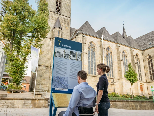 BArrierefreier Stadtrundgang Eine Person im Rollstuhl betrachtet eine Infotafel vor einer Kirche mit gotischem Turm.