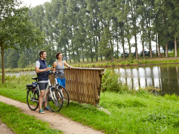 radfahren-lathen-naturpark-h-mmling-44 Zwei Radfahrende machen Pause am Flussufer und blicken lächelnd über das Wasser im Naturpark.