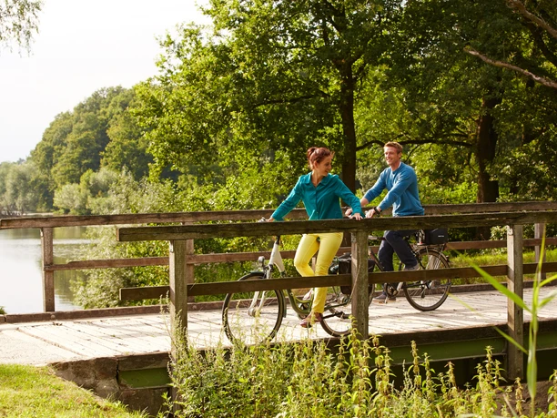 glm1327__3926 Zwei Personen radeln über eine hölzerne Brücke durch eine grüne, sonnige Flusslandschaft.