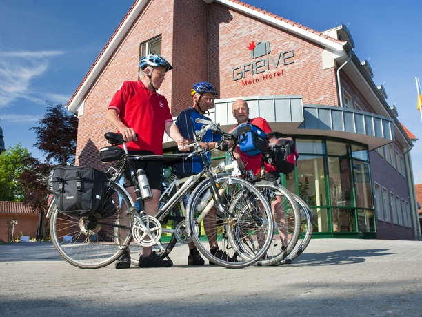 greive-ansicht-radfahrer Drei Radfahrer mit Helmen und Gepäcktaschen vor dem Hotel Greive bei sonnigem Wetter im Emsland.