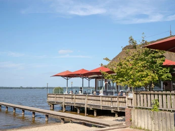 strandcafé Ein gemütliches Strandcafé mit Reetdach direkt am Wasser, umgeben von roten Sonnenschirmen.