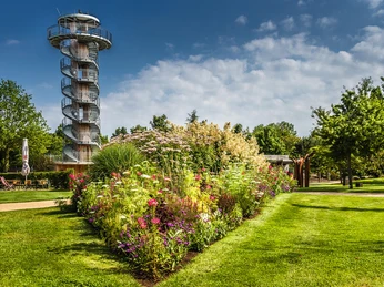 park_der_gaerten Blumenbeet im Park mit bunten Blüten vor einem spiralförmigen Aussichtsturm an einem sonnigen Tag.