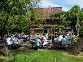 spieker_gaststaette Ein lauschiger Gastgarten vor einem historischen Fachwerkhaus, belebt von geselligen Menschen.