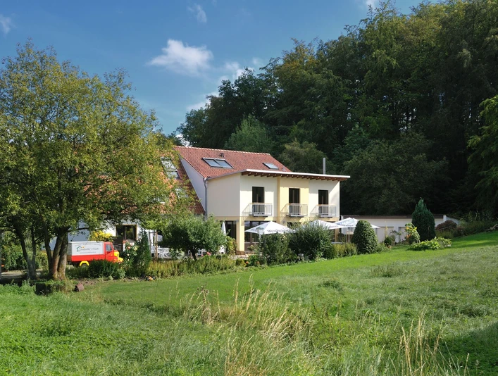 Ein Landhaus mit rotem Dach vor einem Wald, von Wiesen umgeben. Terrasse mit Sonnenschirmen bewohnt.