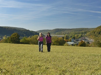 Altenbeken Zwei Frauen wandern über eine Wiese, im Hintergrund das Altenbeker Viadukt und bewaldete Hügel.
