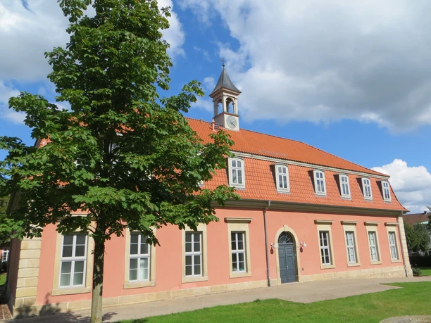 Romantik Bad Rehburg Rosafarbenes Gebäude mit rotem Ziegeldach, Gauben und kleinem Turm, von grünem Baum flankiert.