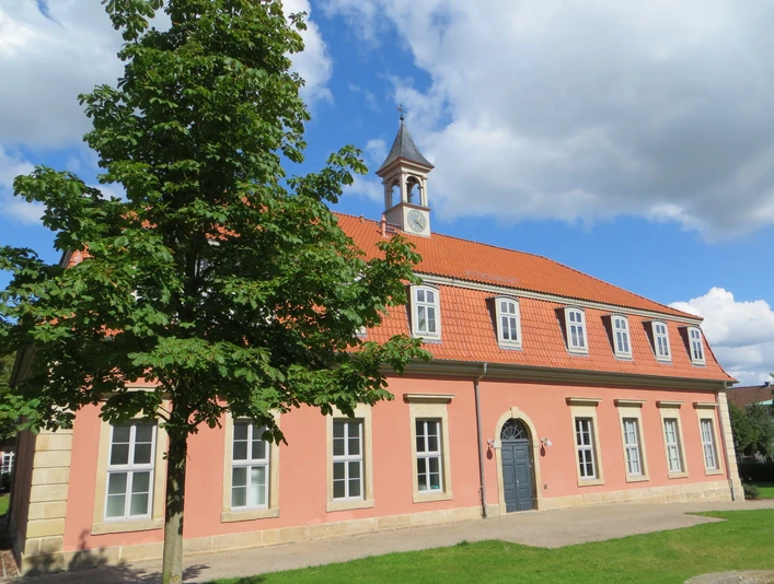 Romantik Bad Rehburg Rosafarbenes Gebäude mit rotem Ziegeldach, Gauben und kleinem Turm, von grünem Baum flankiert.