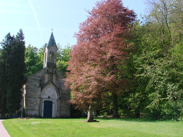 Friederiken-Kapelle Eine historische Kapelle steht neben einem hochgewachsenen Baum in einer grünen Parklandschaft.