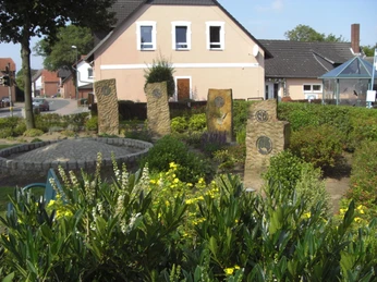 Brunnenplatz Münchehagen Auf einem belebten Platz stehen vier versetzt angeordnete Steinplatten zwischen blühenden Pflanzen.