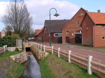 Dorfgraben Winzlar Backsteinbauten mit einem schmalen Wassergraben und bepflastertem Weg in einer ländlichen Szenerie.