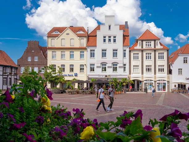 Blick über den Lingener Marktplatz mit historischen Fassaden, blühenden Blumen und Passanten.