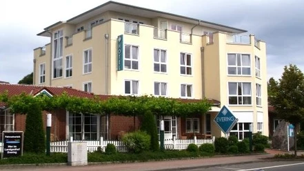 Mehrstöckiges Hotelgebäude mit Restaurantbereich, heller Fassade, begrüntem Eingang und Straßenseite.