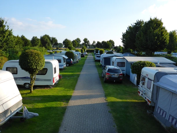 Reihen von Wohnwagen und Autos entlang eines gepflasterten Weges auf gepflegtem, grünen Campingplatz.