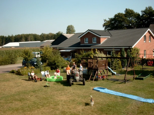 AaSchleife_Spelle_Aussenbereich.png Kinder spielen im Garten eines Landhofs mit Spielgeräten, Planschbecken und kleinem Traktor.