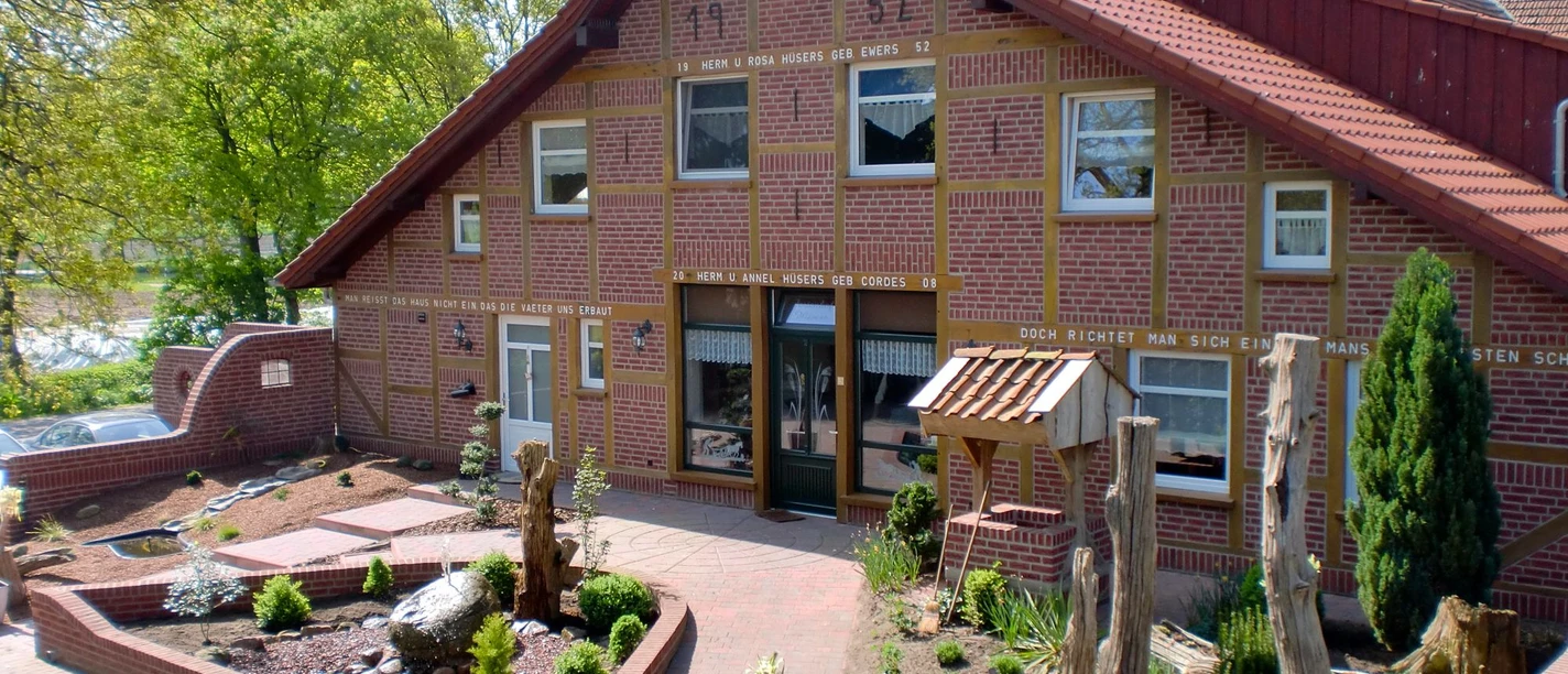 Ferienhof_Huesers__Haren_-_Aussenansicht.jpg Backsteinhaus mit rotem Satteldach, bepflanztem Vorgarten und dekorativen Holzelementen.