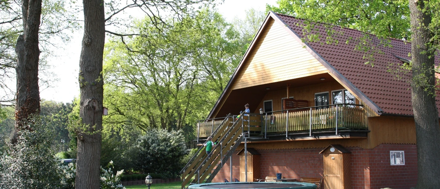Westermann_Herzlake_-_mini.jpg Ferienhofgebäude mit rotem Dach zwischen alten Eichen, davor Wiese mit Trampolin im Sonnenschein.