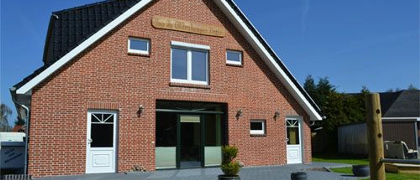 Werlte_-_Ferienwohnung_Too_dae_Oldenkamper_Deele_-_Aussenansicht.jpg Backsteinhaus mit dunklem Dach und zwei Eingängen, umgeben von Rasen und sonnigem Himmel.