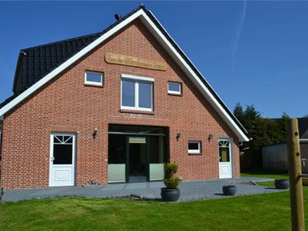 Werlte_-_Ferienwohnung_Too_dae_Oldenkamper_Deele_-_Aussenansicht.jpg Backsteinhaus mit dunklem Dach und zwei Eingängen, umgeben von Rasen und sonnigem Himmel.