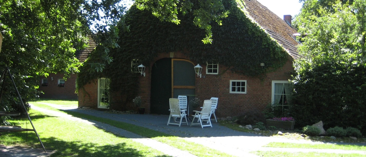 Ferienwohnung_Ottens_Haren4_mini.jpg Backsteingehöft mit Efeubewuchs, Hofeinfahrt und Gartentischgruppe im sonnigen Grünen.
