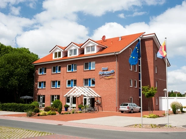 HotelAquamarin_PapenburgHotelansicht.JPG Backsteingebäude mit rotem Dach, blauen Fensterrahmen und Hoteleingang mit Glasvordach in Papenburg