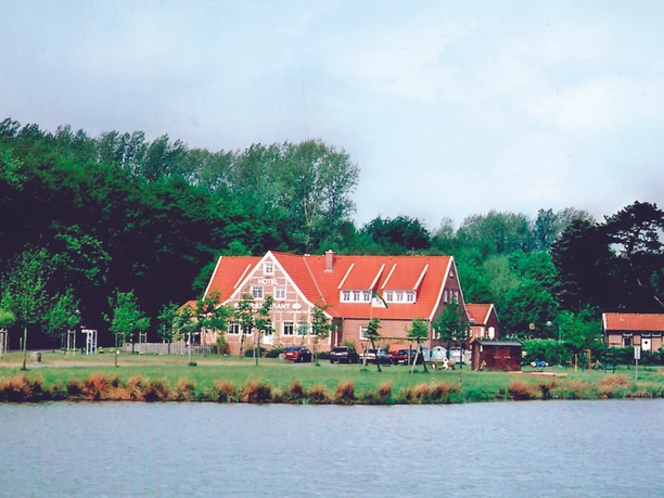 H_LathenerMarsch_Haus_Lathen_mini.jpg Backsteingebäude mit rotem Dach am Seeufer, umgeben von grünen Bäumen und geparkten Autos.