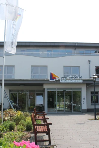 Jugendgaestehaus_Johannesburg_in_Papenburg_-_Haupteingang_klein.jpg Moderner Eingangsbereich des Jugendgästehauses Johannesburg mit Blumenbeeten und Sitzbänken.