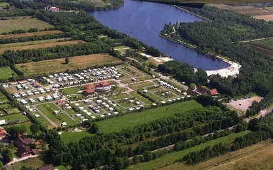 Wiesmoor_Campingplatz_640.jpg Auf dem Bild ist eine Luftaufnahme eines weitläufigen Campingplatzes in Wiesmoor zu sehen, der von üppigem Grün und einem malerischen See umgeben ist. Der Platz ist in rechteckigen Parzellen organisiert, in denen zahlreiche Wohnwagen und Zelte aufgestellt sind. In der Mitte befindet sich ein Zentralgebäude mit einem roten Dach. Der See liegt rechts im Bild und bietet einen sandigen Strandbereich.