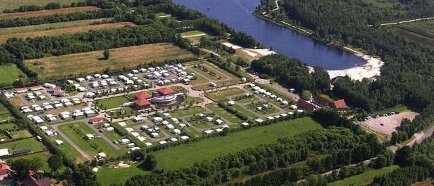 Wiesmoor_Campingplatz_640.jpg Auf dem Bild ist eine Luftaufnahme eines weitläufigen Campingplatzes in Wiesmoor zu sehen, der von üppigem Grün und einem malerischen See umgeben ist. Der Platz ist in rechteckigen Parzellen organisiert, in denen zahlreiche Wohnwagen und Zelte aufgestellt sind. In der Mitte befindet sich ein Zentralgebäude mit einem roten Dach. Der See liegt rechts im Bild und bietet einen sandigen Strandbereich.
