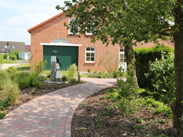 Camping am Deich in Krummhörn-Upleward Backsteingebäude am Deich mit grünem Tor, bepflanztem Gartenweg und Bäumen im Vordergrund