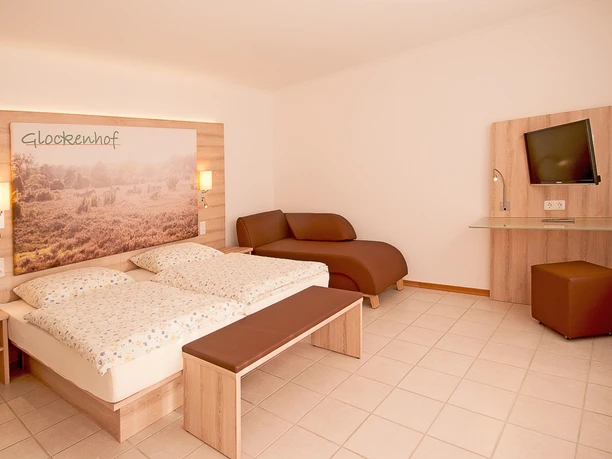 Schlafzimmer Helles Hotelzimmer mit Doppelbett, Wandbild vom Glockenhof, brauner Liege und Schreibtisch.