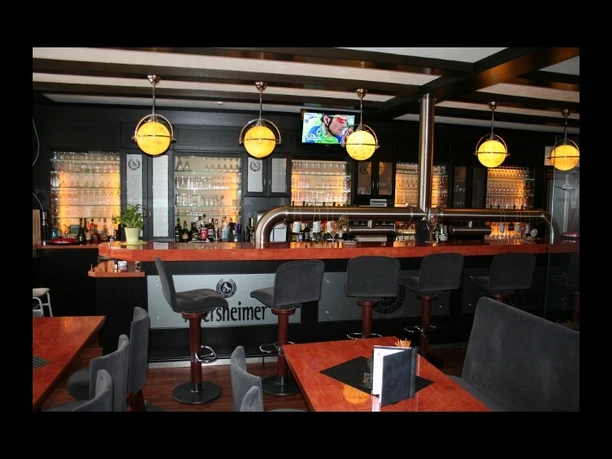 hotel-corveyerhof-bierstube Eine Bierstube mit moderner Bar, gepolsterten Barhockern und hölzernen Tischen, beleuchtet durch mehrere runde Pendelleuchten.