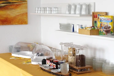 pension-zurweser-fruehstueck Frühstücksbuffet mit Cerealien, Marmelade und Saft, vor warmen Erdfarben und schlichtem Geschirr.