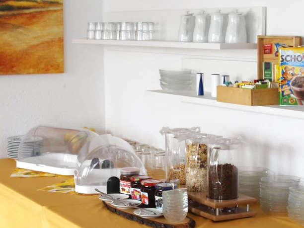 pension-zurweser-fruehstueck Frühstücksbuffet mit Cerealien, Marmelade und Saft, vor warmen Erdfarben und schlichtem Geschirr.