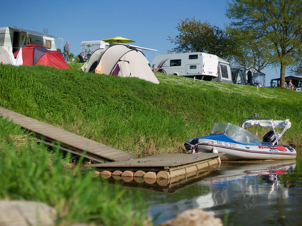 Campingplatz mit Zelten und Wohnwagen am Fluss. Ein Boot ist an einem Steg im Wasser vertäut.