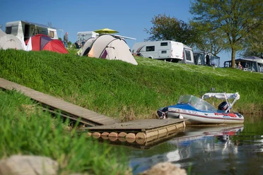 wesercamping-bootsanleger Campingplatz mit Zelten und Wohnwagen am Fluss. Ein Boot ist an einem Steg im Wasser vertäut.