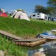 wesercamping-bootsanleger Campingplatz mit Zelten und Wohnwagen am Fluss. Ein Boot ist an einem Steg im Wasser vertäut.