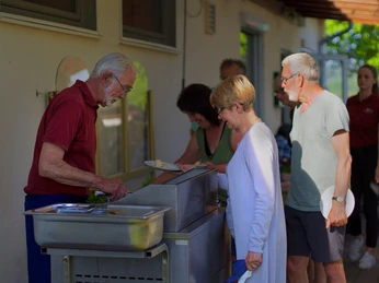 Mehrere Menschen stehen an einer Grillstation, während ein älterer Mann am Grill Fleisch zubereitet.