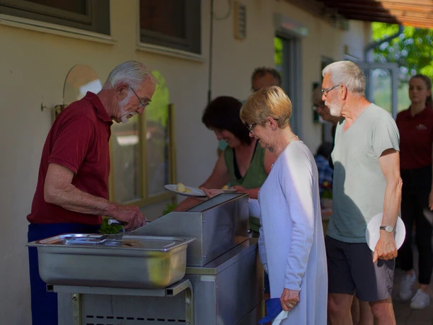 wesercamping-grillabend Mehrere Menschen stehen an einer Grillstation, während ein älterer Mann am Grill Fleisch zubereitet.