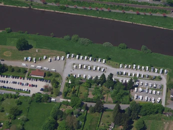 Luftaufnahme eines Wohnmobil-Stellplatzes am Fluss mit geparkten Fahrzeugen und angrenzenden Grünflächen.
