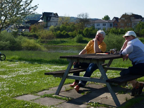 wohnmobil-flossplatz-sitzgruppe Älteres Paar genießt entspannt die Aussicht an einem Holztisch im Grünen mit verstreuten Gänseblümchen.