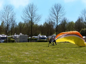Paragleiter startet auf einer grünen Wiese, im Hintergrund stehen Wohnmobile unter Bäumen.