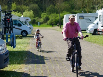 Ein Mann und ein Kind fahren auf einem Fahrradweg neben geparkten Wohnmobilen in einer grünen Umgebung.